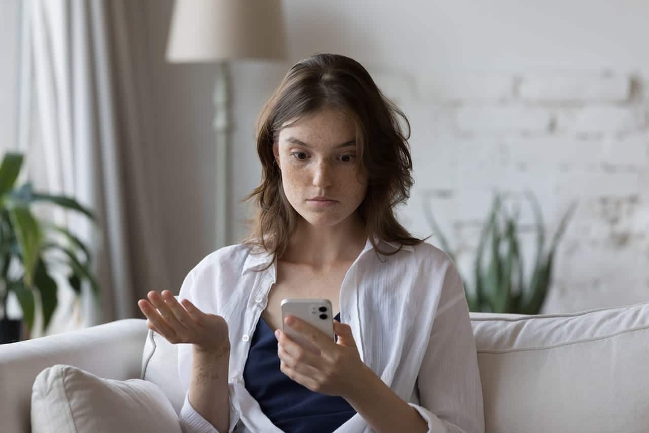 Mulher sentada em um sofá olhando com expressão de choque para o celular que segura