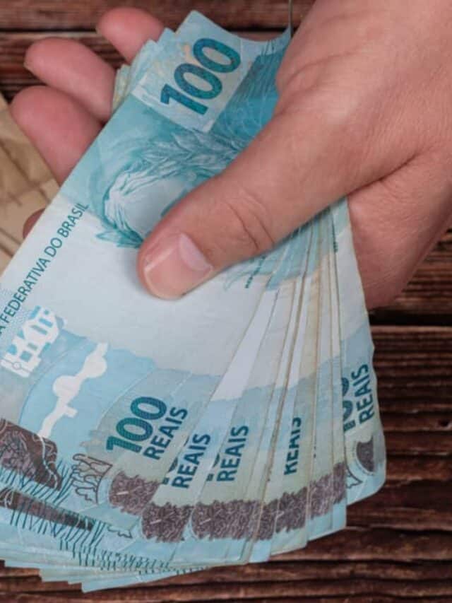 Mão segurando várias notas de 100 reais. Em baixo, sobre uma mesa, há várias notas de 50 reais