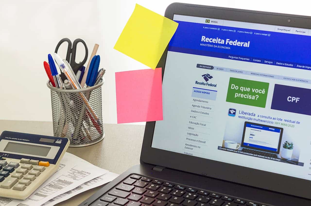 tela de notebook mostra site da Receita Federal. Em cima da mesa tem ainda porta canetas, calculadora e papéis.