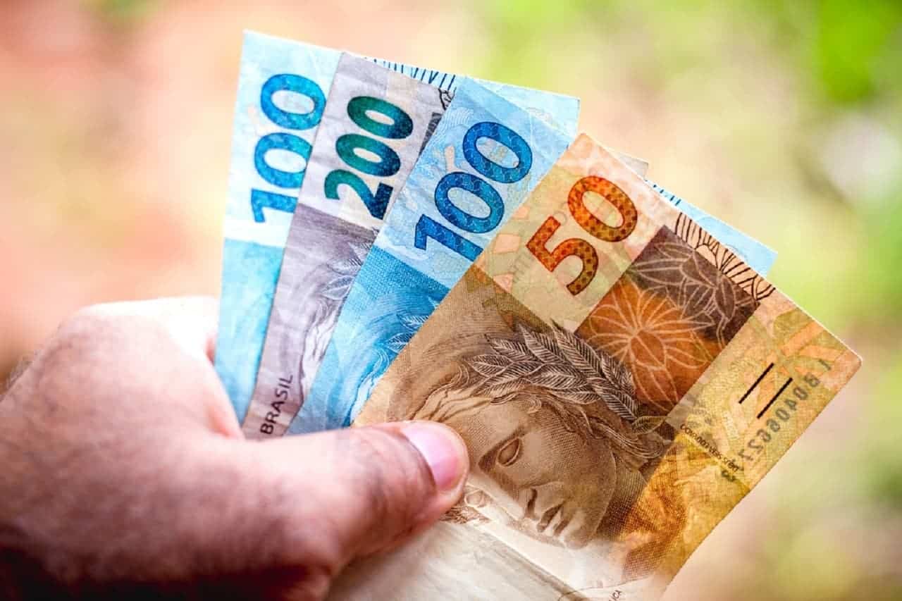 Mão segurando notas de dinheiro de R$50, R$100 e R$200. Fundo desfocado valores a receber dinheiro esquecido