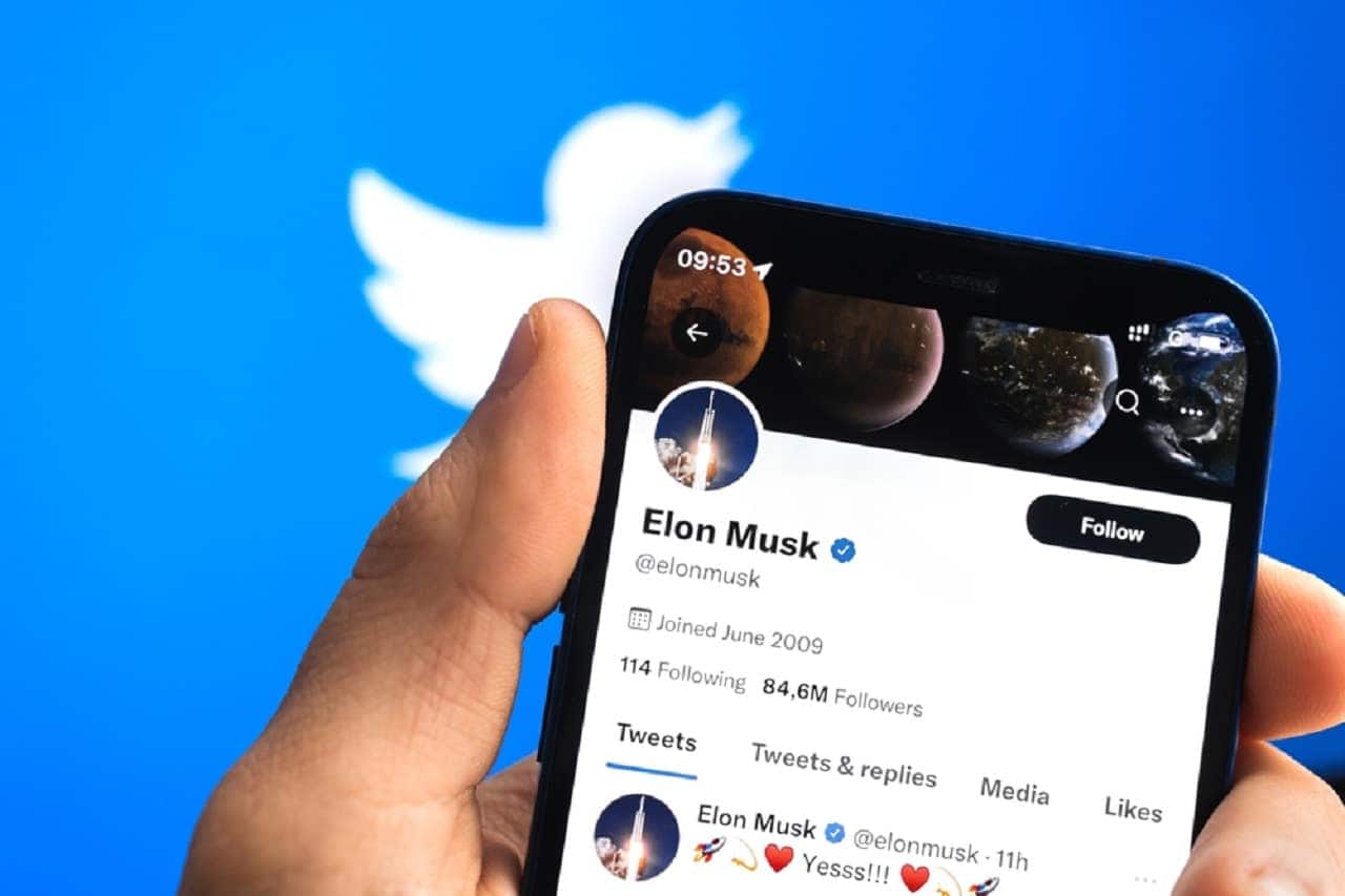 Imagem de uma mãe segurando um celular, com o perfil de Elon Musk no Twitter aberto. Ao fundo, o logo da rede social.