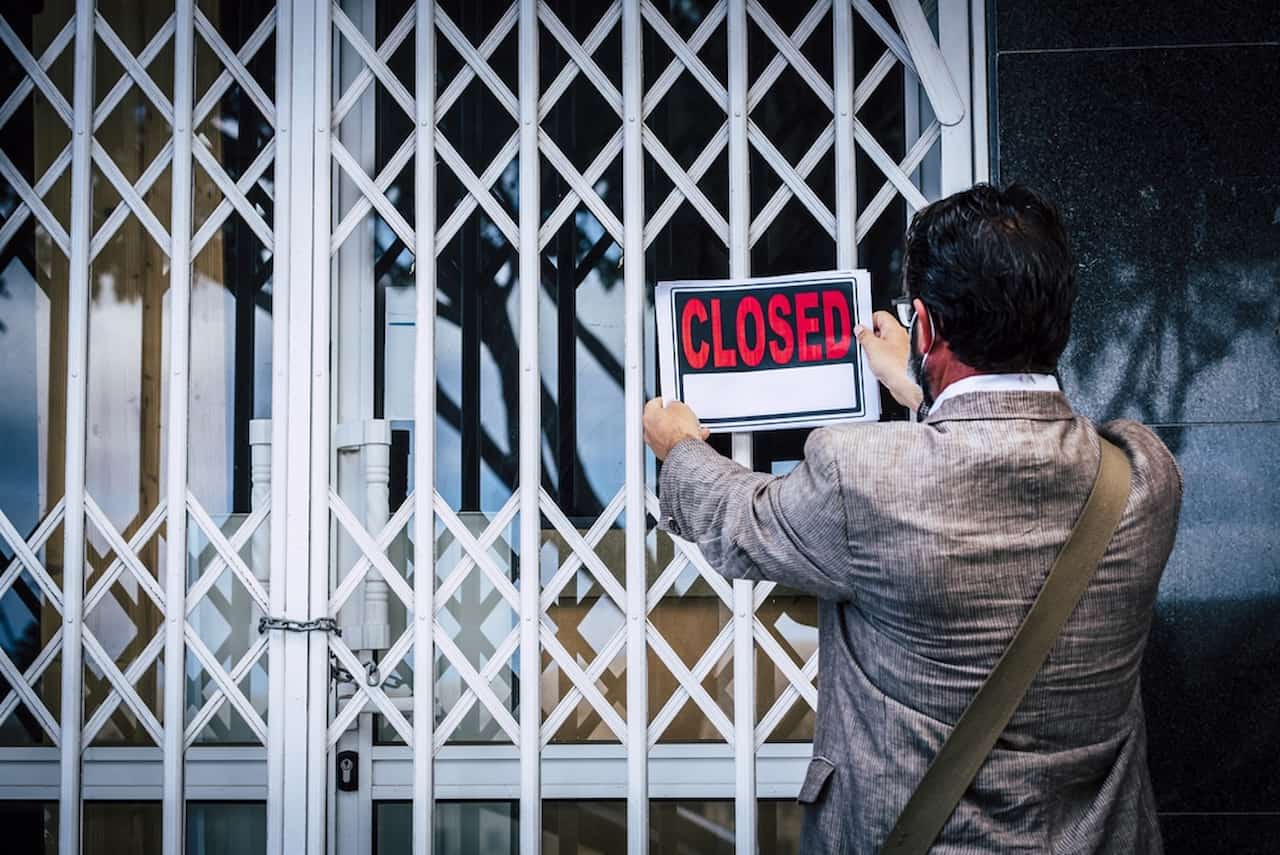 Empresa com as portas fechadas enquanto homem coloca placa com a palavra "Closed", que em português quer dizer "fechado", em uma grade
