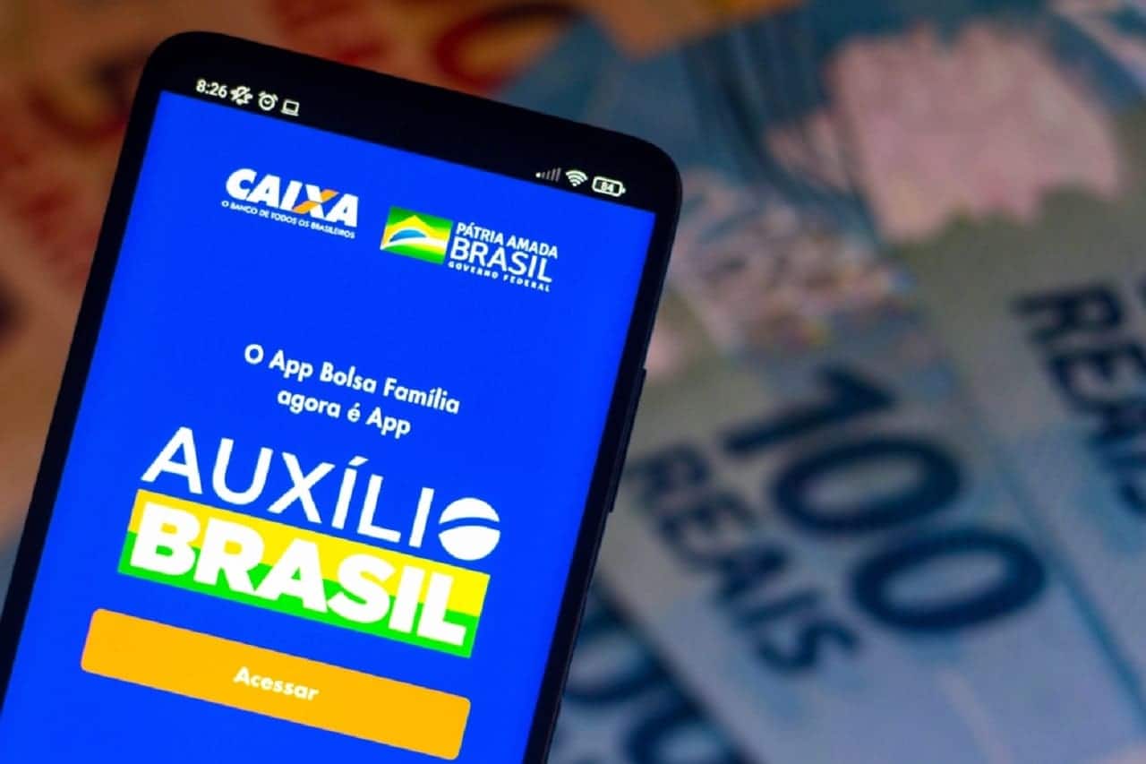 Imagem de um celular com o aplicativo do auxílio brasil aberto. Ao fundo, algumas notas de R$100 desfocadas.