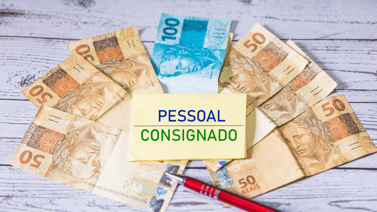 Imagem mota notas de dinheiro de 50 ou 100, com uma placa escrito "pessoal consignado" no meio e uma caneta vermelha e prata em baixo.