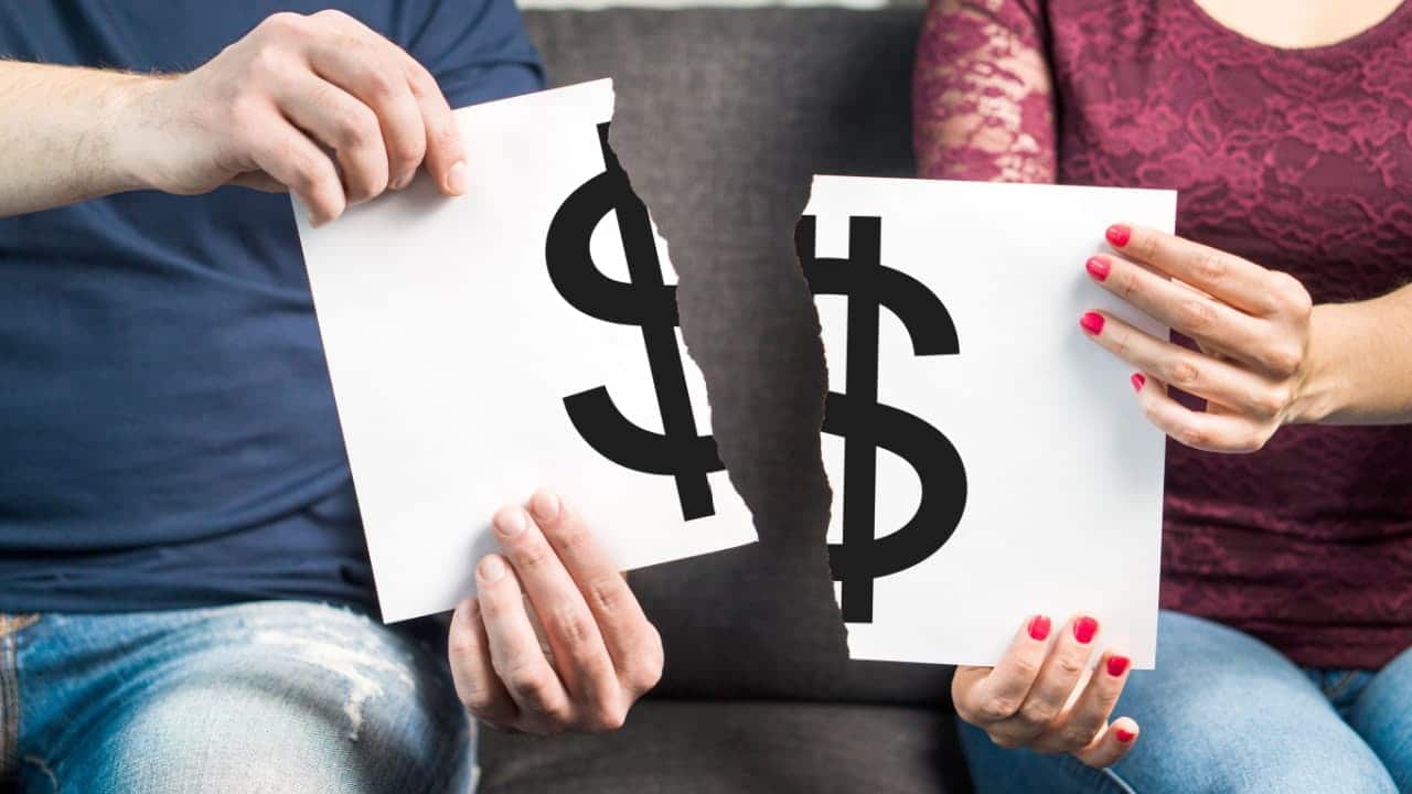 Homem e mulher sentados em um sofá, cada um segurando metade de um papel com cifrão de dinheiro rasgado no meio.