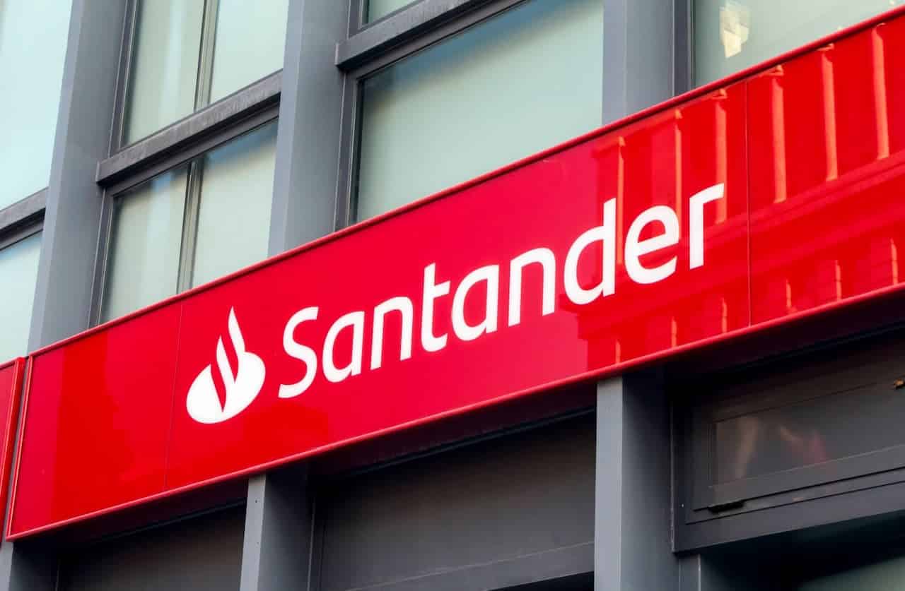 Fachada de uma agência do banco Santander com o letreiro branco sobre a faixa vermelha em um prédio com pilares azuis