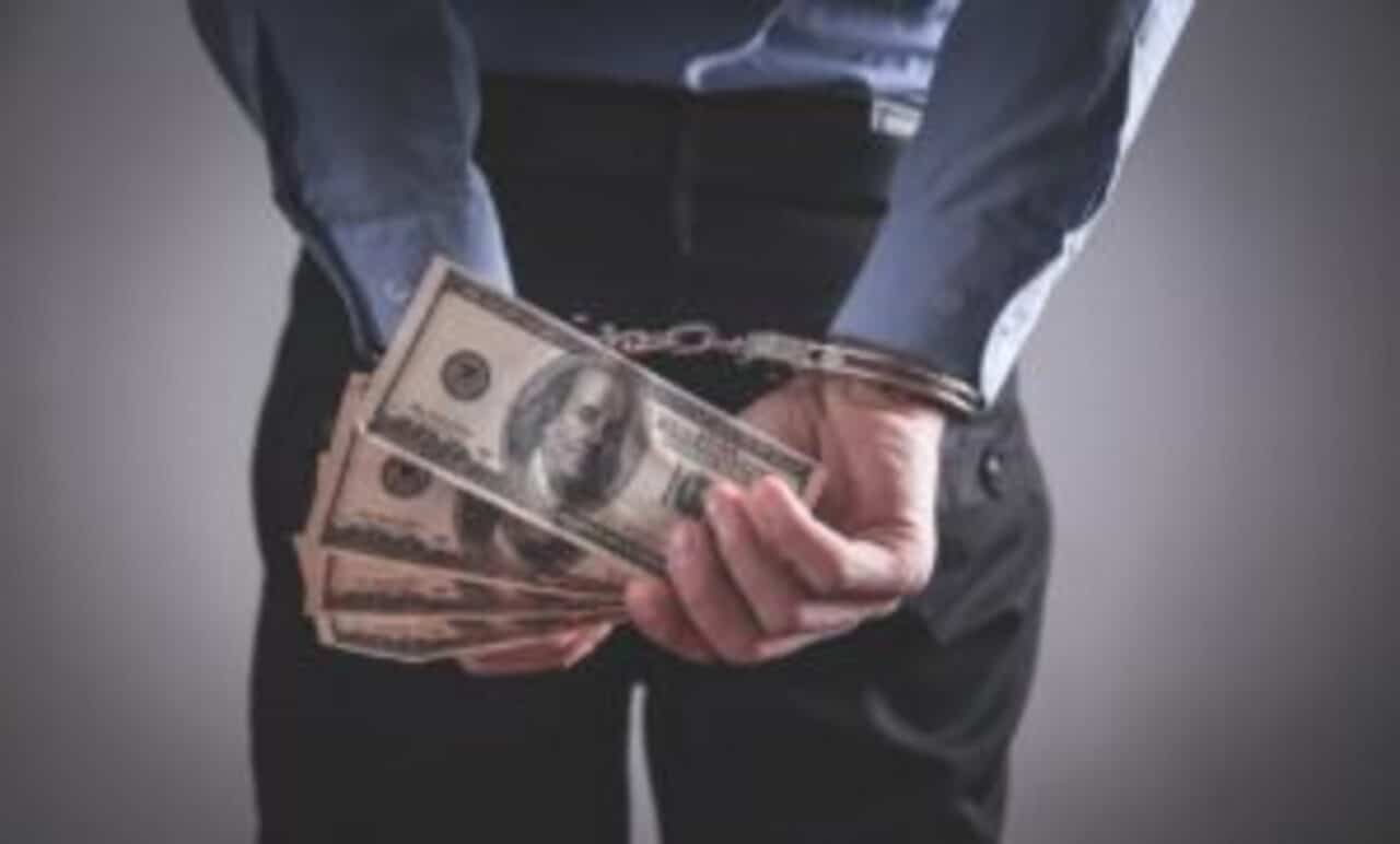 Pessoa com mãos algemadas nas costas e segurando notas de dólar