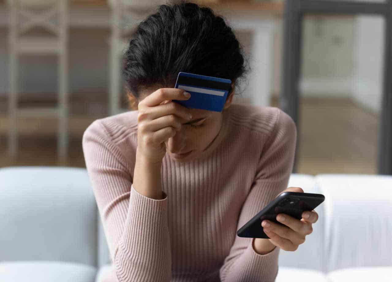 mulher cabisbaixa com cartão em uma mão e celular em outro ao descobrir golpe financeiro
