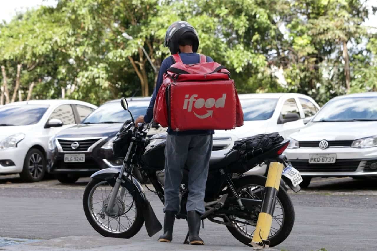 Imagem de um homem de costas com uma mochila de entrega do iFood. Está ao lado de uma moto. Ao fundo, vários carros.
