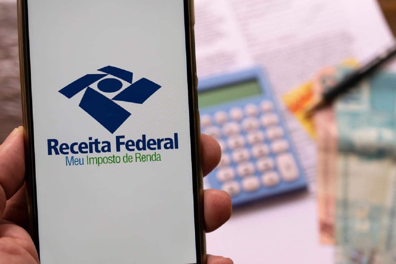 Imagem de uma mão segurando um celular com o aplicativo da Receita Federal, chamado "Meu Imposto de Renda". Ao fundo, documentos, dinheiro e uma calculadora.