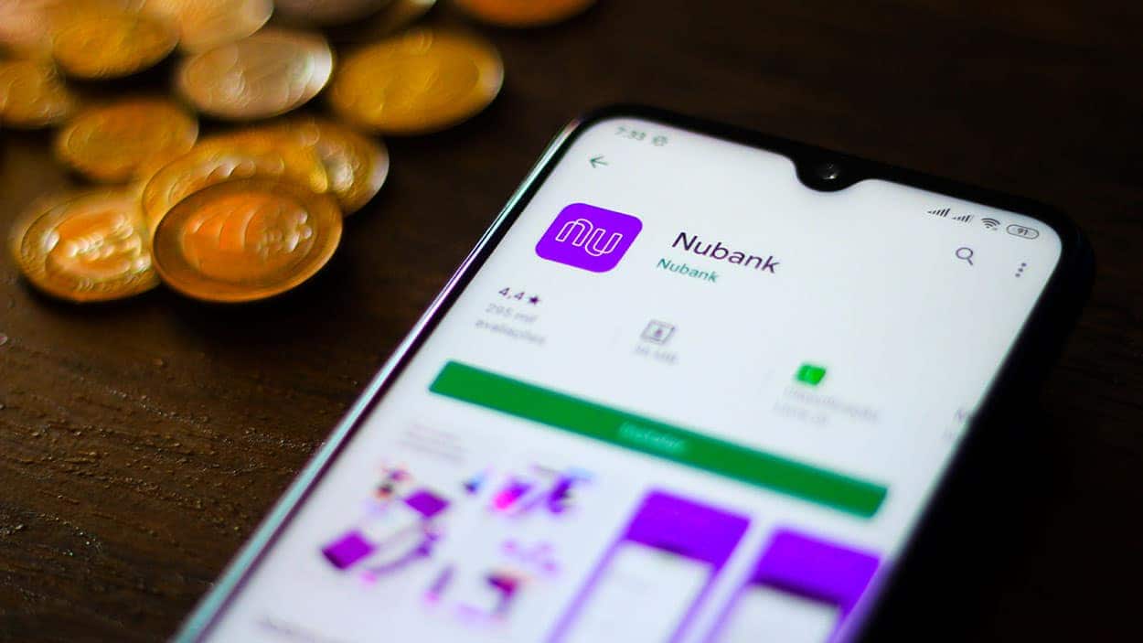 Imagem de um celular com a tela ligada no app do Nubank, ao lado de algumas criptomoedas