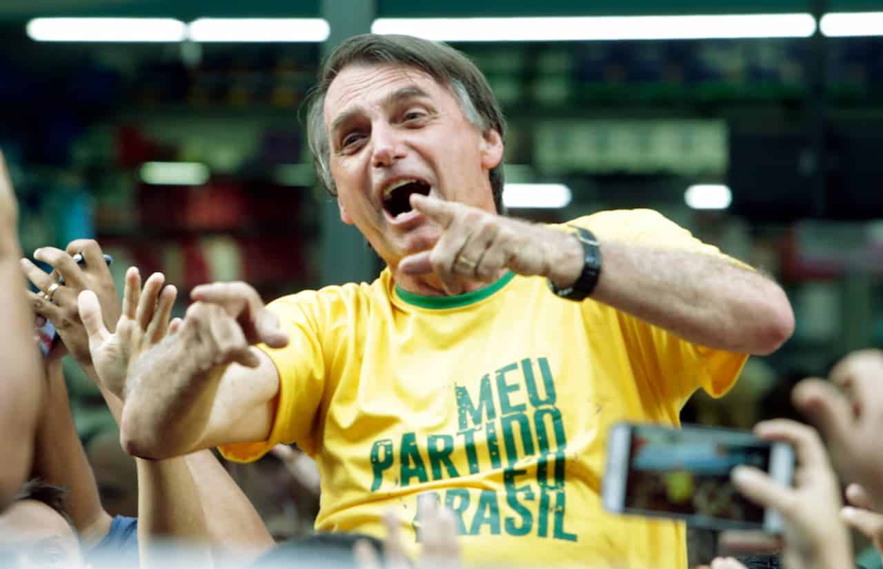 Imagem do ex-presidente, Jair Bolsonaro, com camisa verde e amarela, sorrindo e apontando os dedos para frente, durante comício eleitoral.