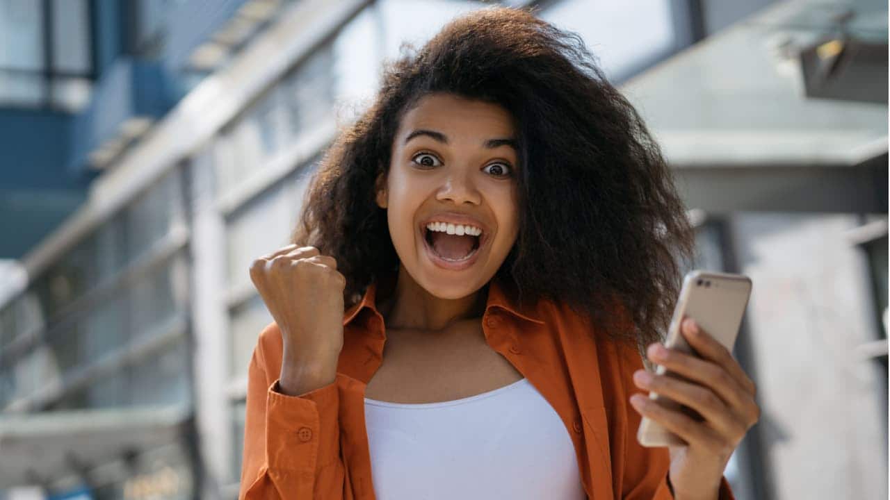 Mulher feliz em ambiente aberto comemorando com um celular em uma das mãos