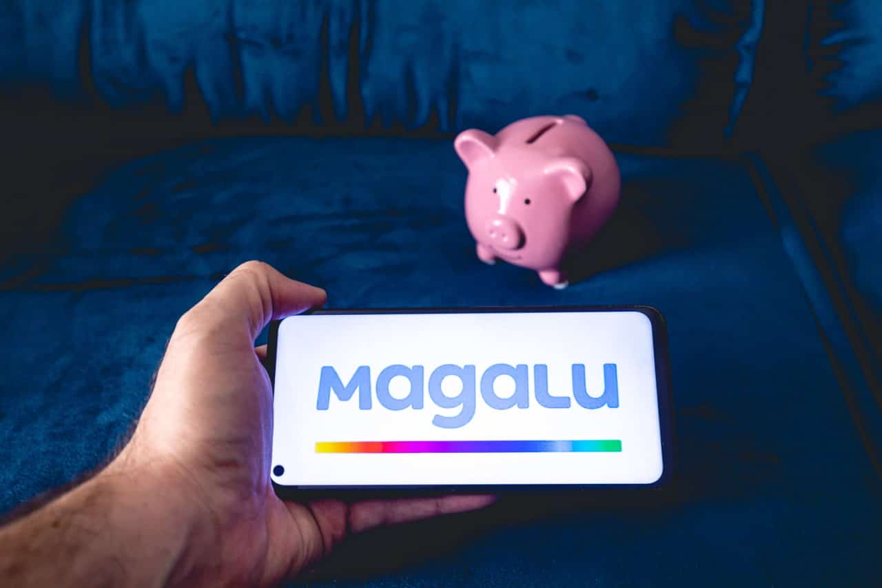 Mão segurando celular que mostra logo do Magalu (Magazine Luiza) em primeiro plano. Ao fundo, um cofrinho rosa em formato de porco.