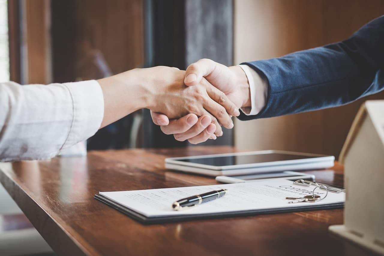 Duas pessoas se cumprimentando, como que firmando um acordo. Ha uma mesa com papeis e caneta embaixo das mãos