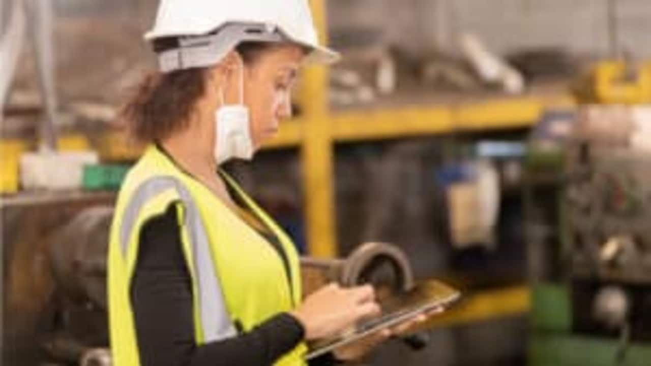 Mulher de perfil vestindo EPIs mexendo em um tablet. Ferramentas desfocadas ao fundo