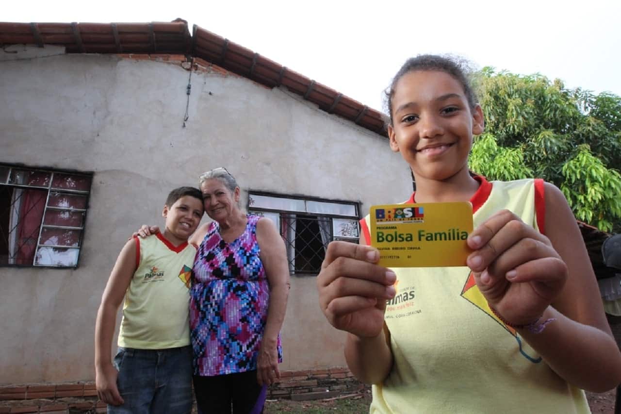 Imagem de uma família, em frente a uma casa simples, composta por uma senhora, um menino e uma menina. A menina segura o cartão do Bolsa Família e sorri.