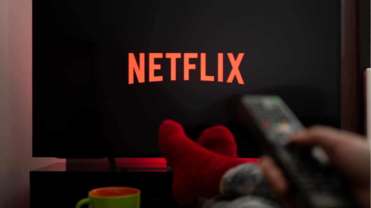 Pés de uma pessoa deitada segurando controle remoto apontado para uma TV. A tela do aparelho mostra o logo da Netflix.