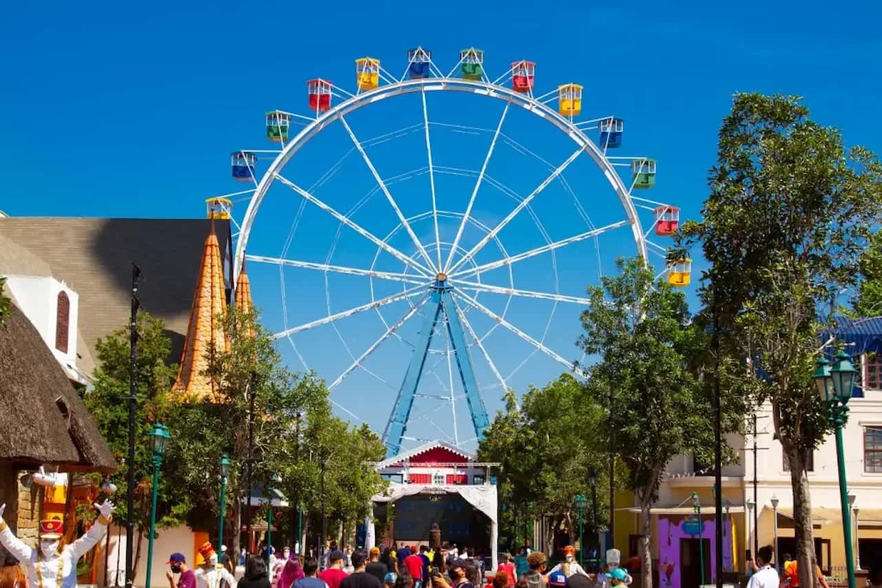 Roda gigante do Hopi Hari ao centro, muitas pessoas circulando pelo parque em meio a árvores e casas