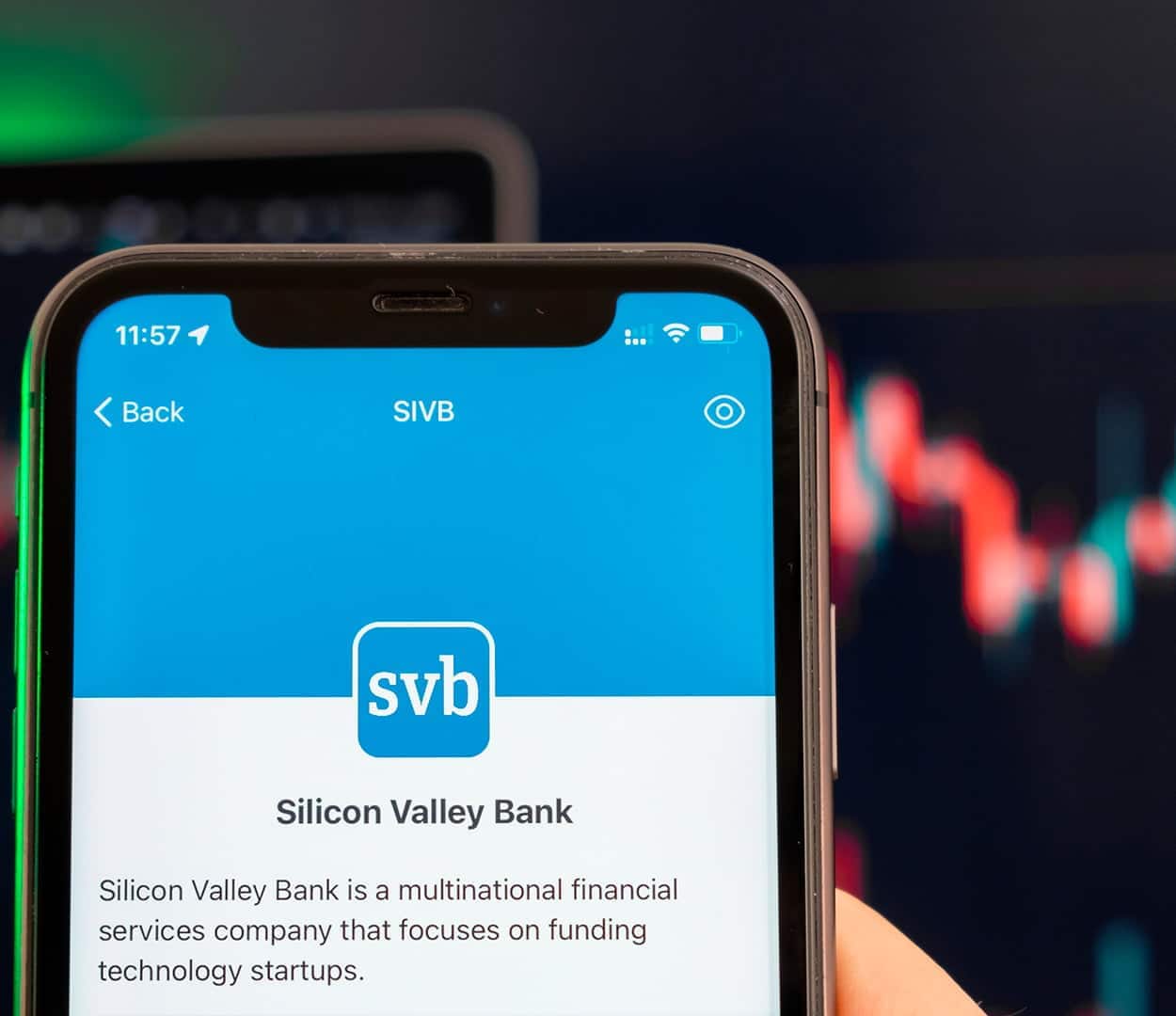 Imagem de um celular com a tela focada no logo do SVB. Ao fundo, um gráfico com movimento de baixa.