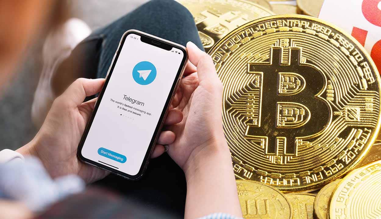Mãos de uma pessoa segurando celular que mostra tela de login do aplicativo Telegram. Ao lado, outra imagem mostra uma moeda que representa o criptoativo Bitcoin.