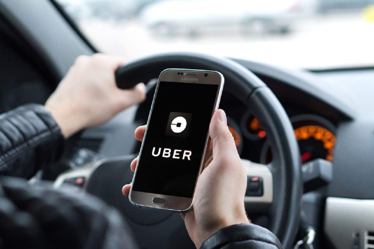 Imagem de um motorista, segurando o volante com uma mão. Na outra mão, um celular com o aplicativo da Uber aberto.
