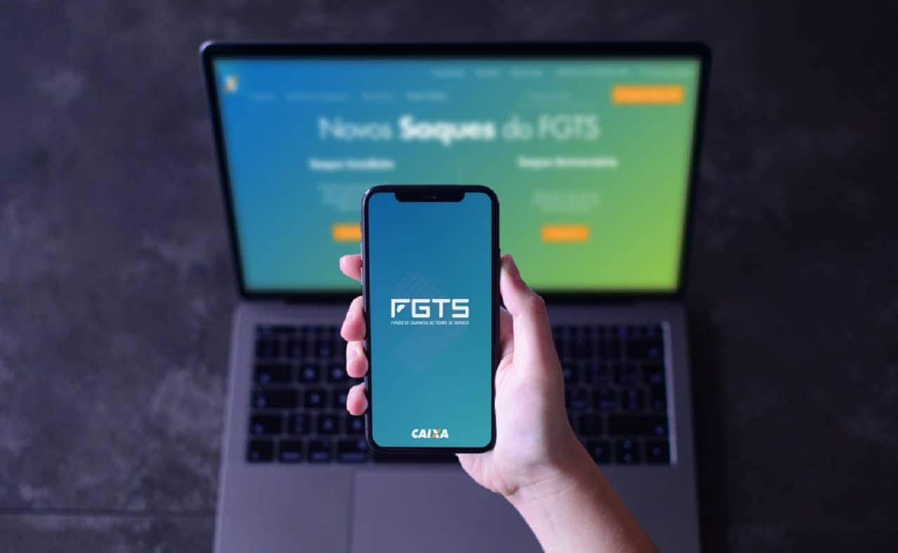 Imagem de uma mão segurando um celular com o aplicativo do FGTS aberto. Ao fundo, um computador desfocado com o site do FGTS aberto.