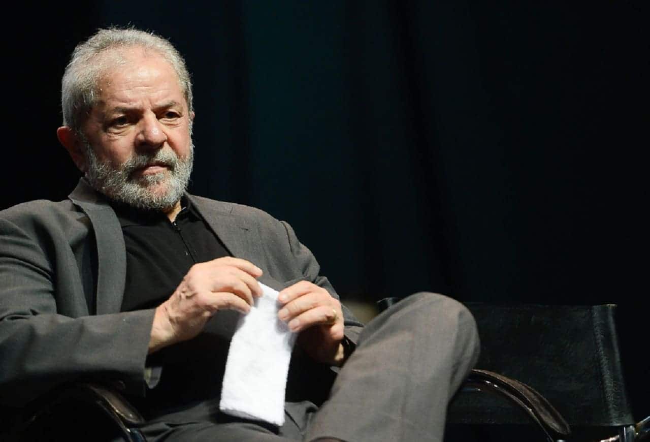 Presidente Lula sentado em cadeira com a perna esquerda sobre a perna direita. Segura as mãos um papel branco. O fundo é todo escuro.