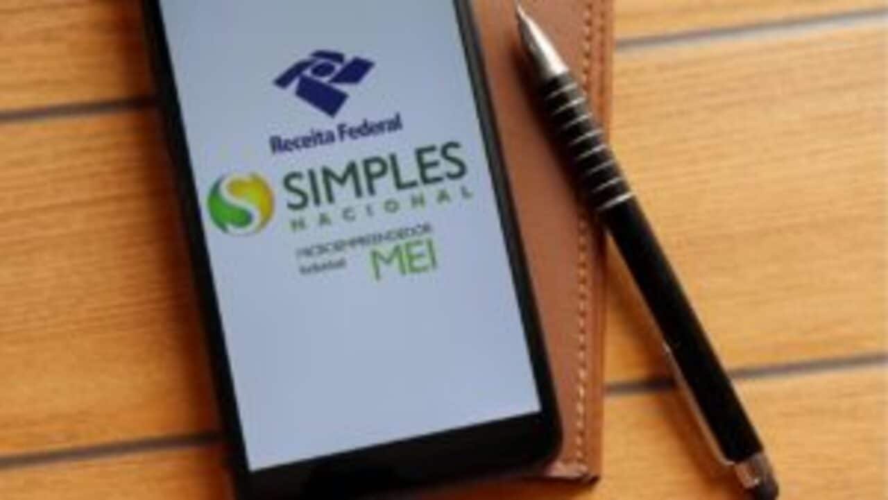 Uma caneta e um celular que exibe logo do MEI, Simples Nacional e Receita Federal em fundo branco, ambos estão posicionados em cima de uma carteira da cor marrom