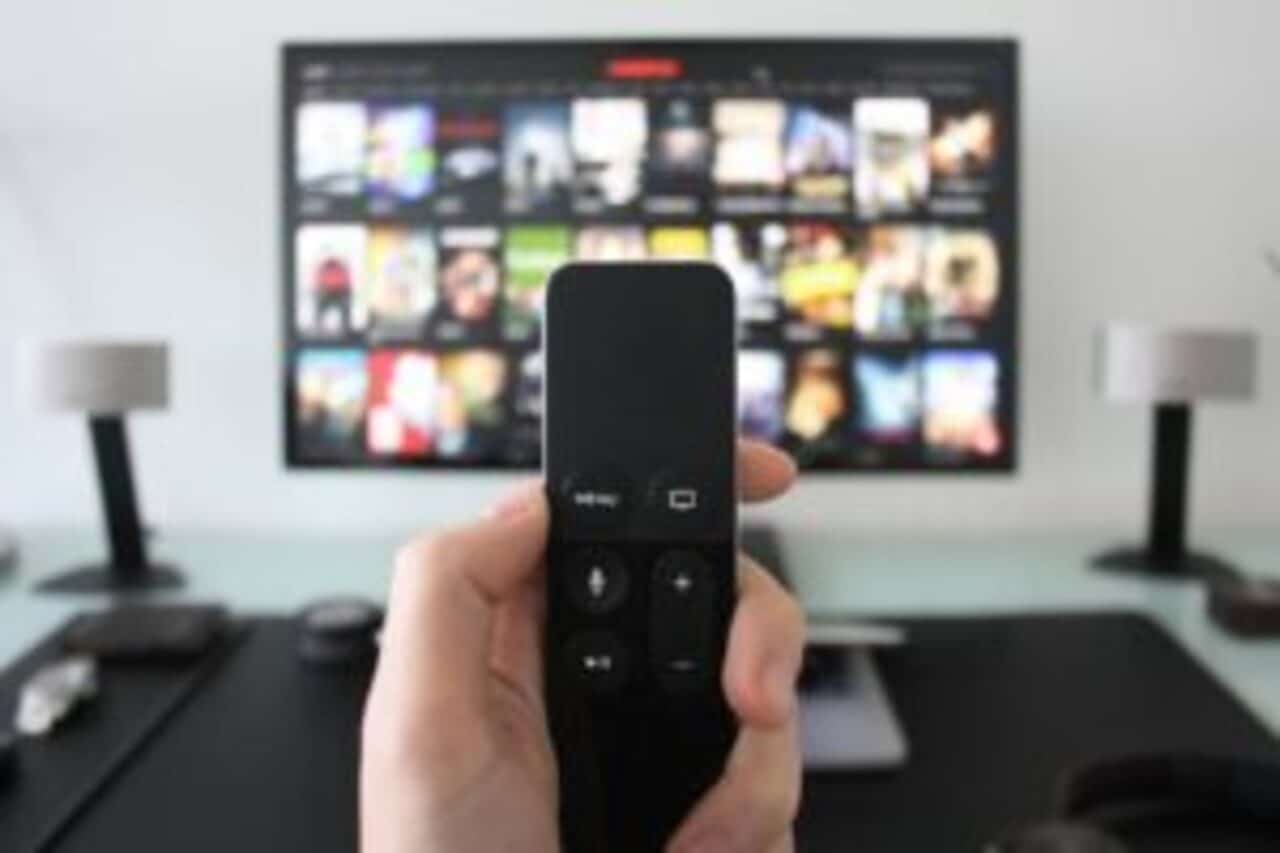 Mão segurando controle remoto e plataformas de streaming ao fundo