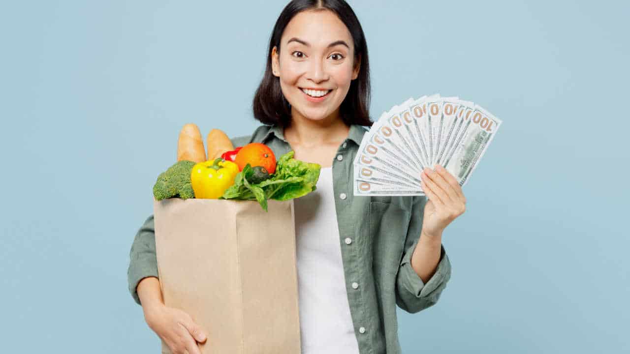 mulher segurando um pacote de alimentos em uma mão e, na outra, um “leque” formado com dinheiro
