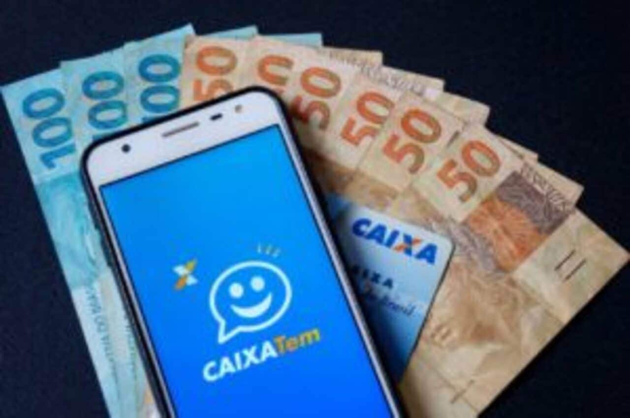 Celular exibindo logotipo do aplicativo Caixa Tem sobre várias notas de 100 e 50 reais.