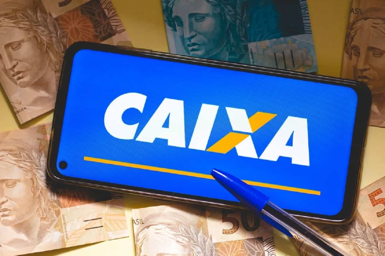Celular posicionado na horizontal que mostra logotipo da Caixa. O aparelho está ao lado de uma caneta e sobre várias notas de real.