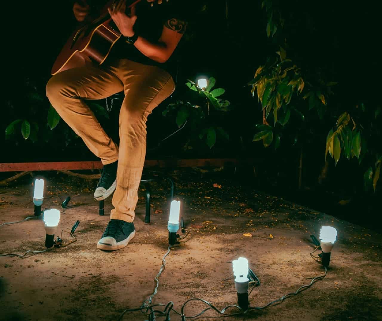 Imagem do corpo de um cantor sertanejo, tocando violão, sentado em um banco. Está em um ambiente com plantas e iluminação baixa.