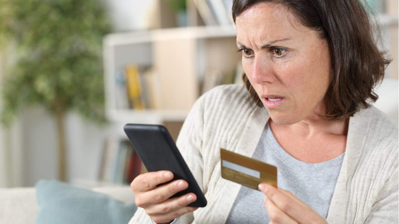 Mulher olhando para o celular com expressão de choque e cartão de crédito na mão