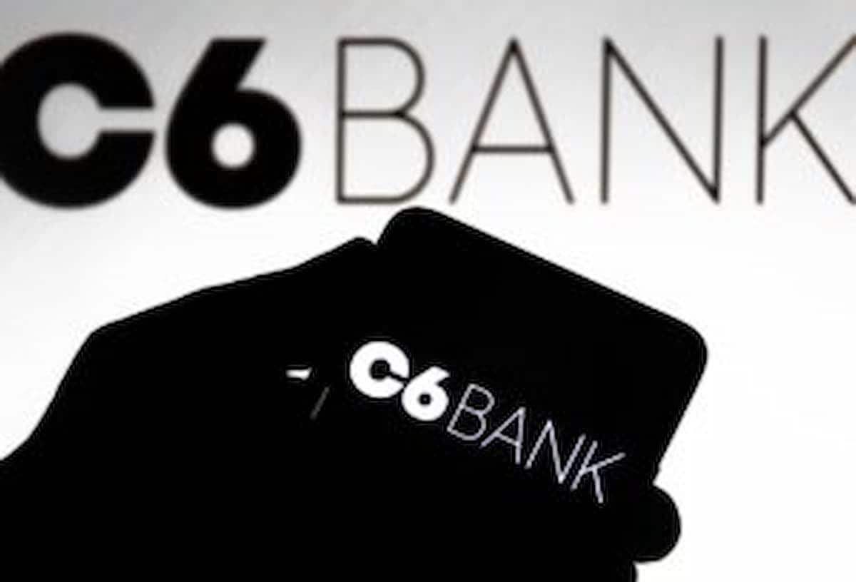Sombra de uma mão segurando um celular que exibe logo do C6 Bank. No fundo da imagem, também aparece o nome do banco digital.