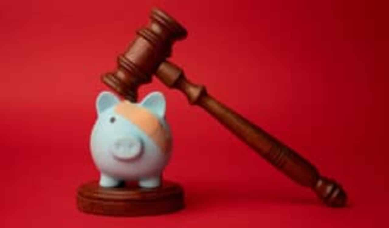Malhete batendo em um porquinho, que representa as finanças. O objeto tem um curativo em um dos olhos. O fundo da imagem está em vermelho.