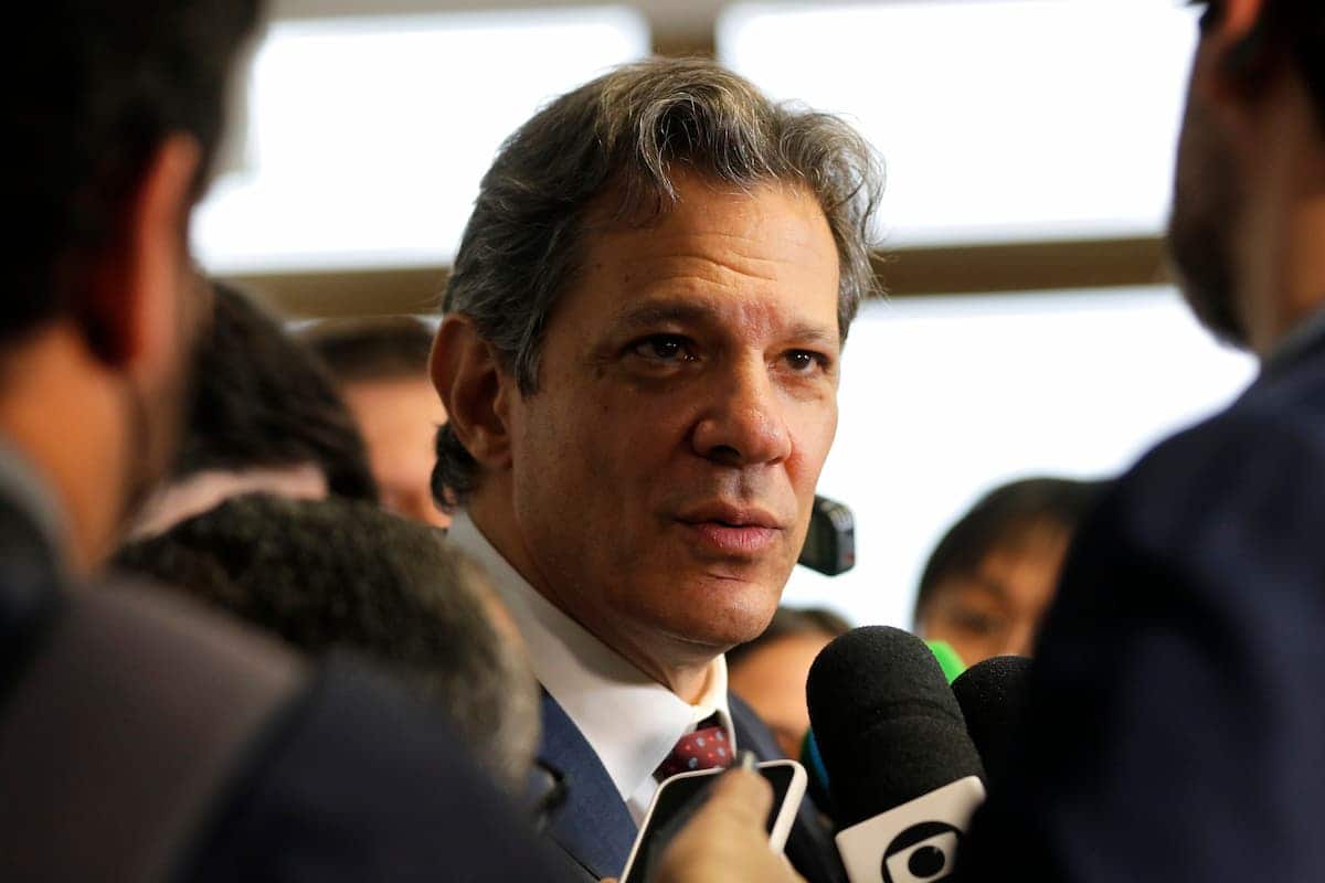 Ministro da Fazenda Fernando Haddad sendo entrevistado, com um microfone e um celular, de pessoas ao seu redor, perto de seu rosto iof