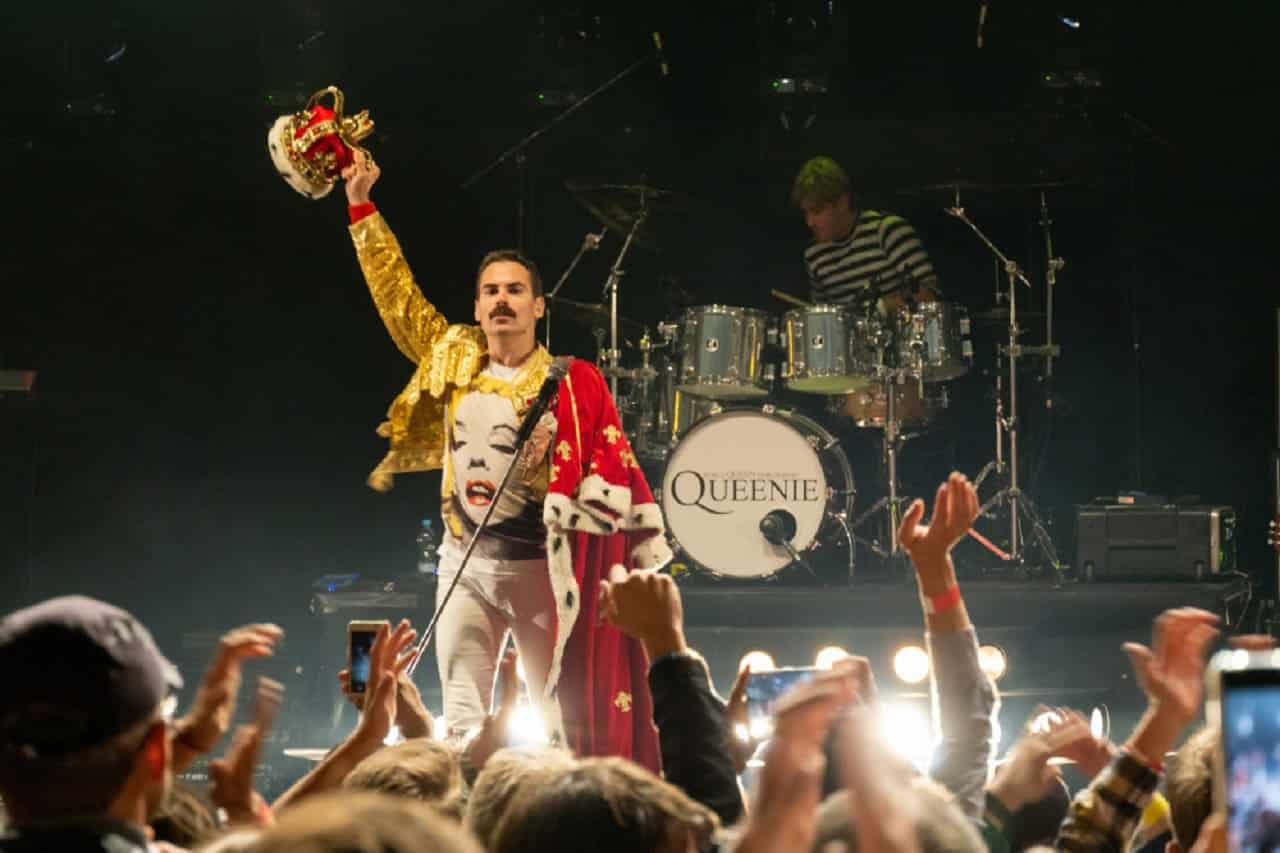Freddie Mercury em um palco, acompanhado do resto da banda Queen. Em sua frente, o público do show.