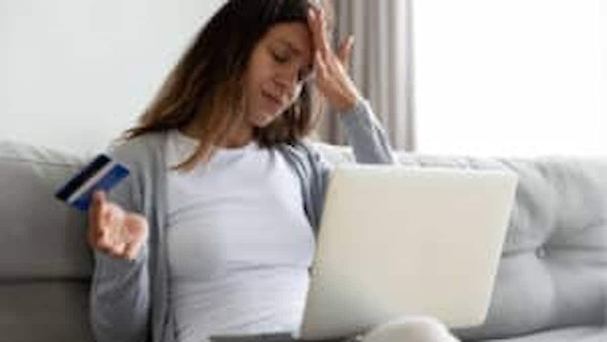 Mulher lamenta com a mão na testa, segurando cartão de crédito e com notebook no colo