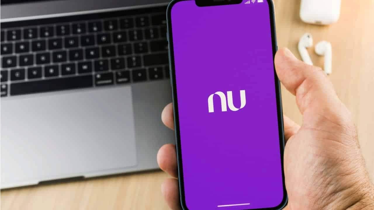 Pessoa segurando celular com app Nubank aberto. Em segundo plano, um notebook e um fone de ouvido