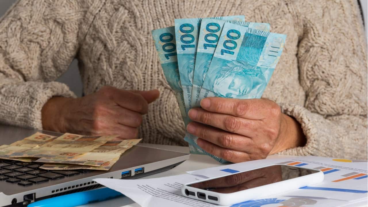 Perto de uma mesa com com contas, celular e notebook, uma pessoa segura notas 4 notas de 100 reais