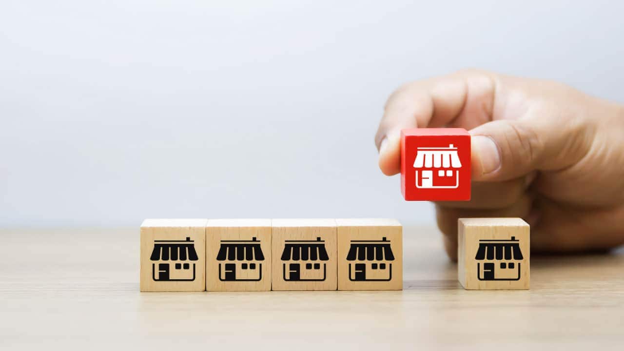 Sequência de cubos de madeira sobre uma mesa, lado a lado, ilustrados com fachadas de lojas. Acima dos objetos, uma mão segura um cubo quase idêntico, mas em cor de destaque, em movimento para encaixá-lo na mesma sequência de itens.