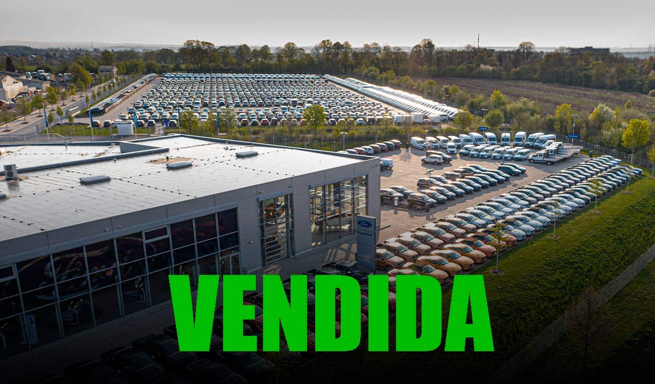 Imagem de um pátio cheio de carros em frente a um grande galpão. O galpão pode ser identificado por um logo da Ford que está discreto na foto. Na parte de baixo da imagem, centralizado, está a palavra "Vendida"