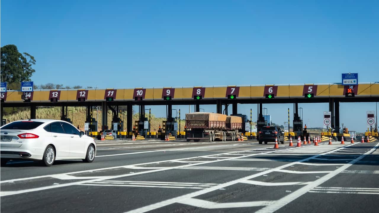 Imagem de uma rodovia na parte de um pedágio, há um caminhão e um carro branco em destaque, mas há outros poucos veiculos