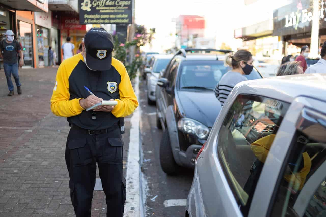 Pessoa uniformizada parada ao lado de um carro, aplicando multa de trânsito