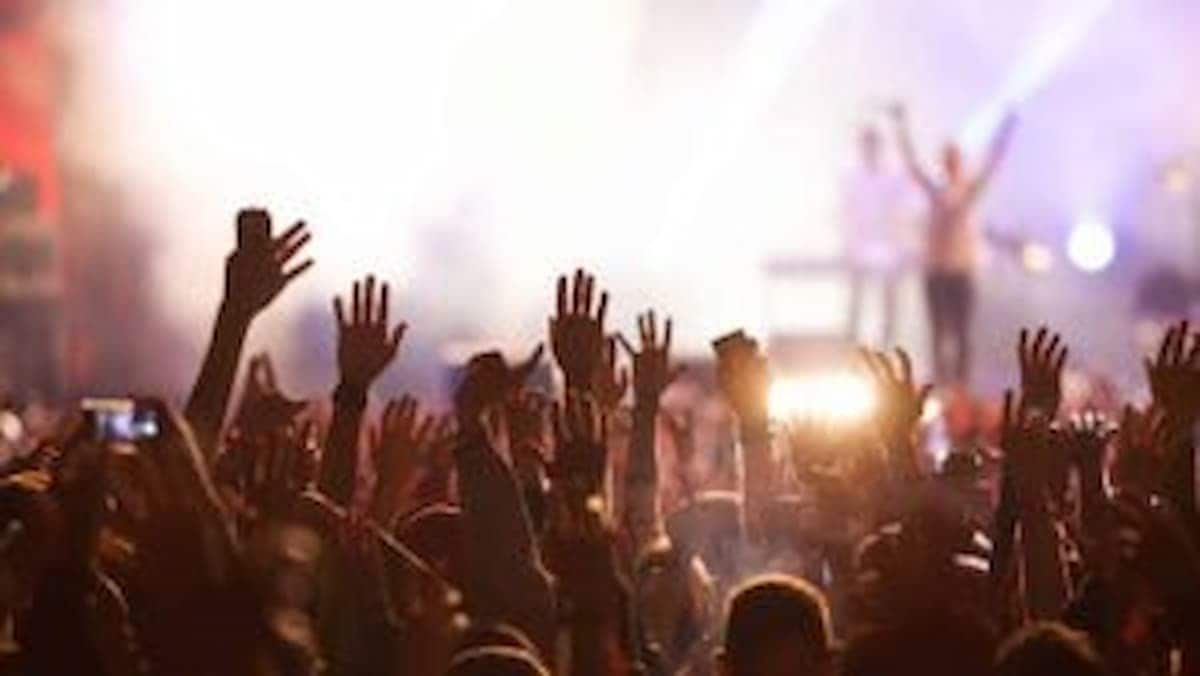 Imagem de um show ou festa com várias pessoas em primeiro plano, com mãos erguidas, e um palco com alguém em cima ao fundo, desfocado