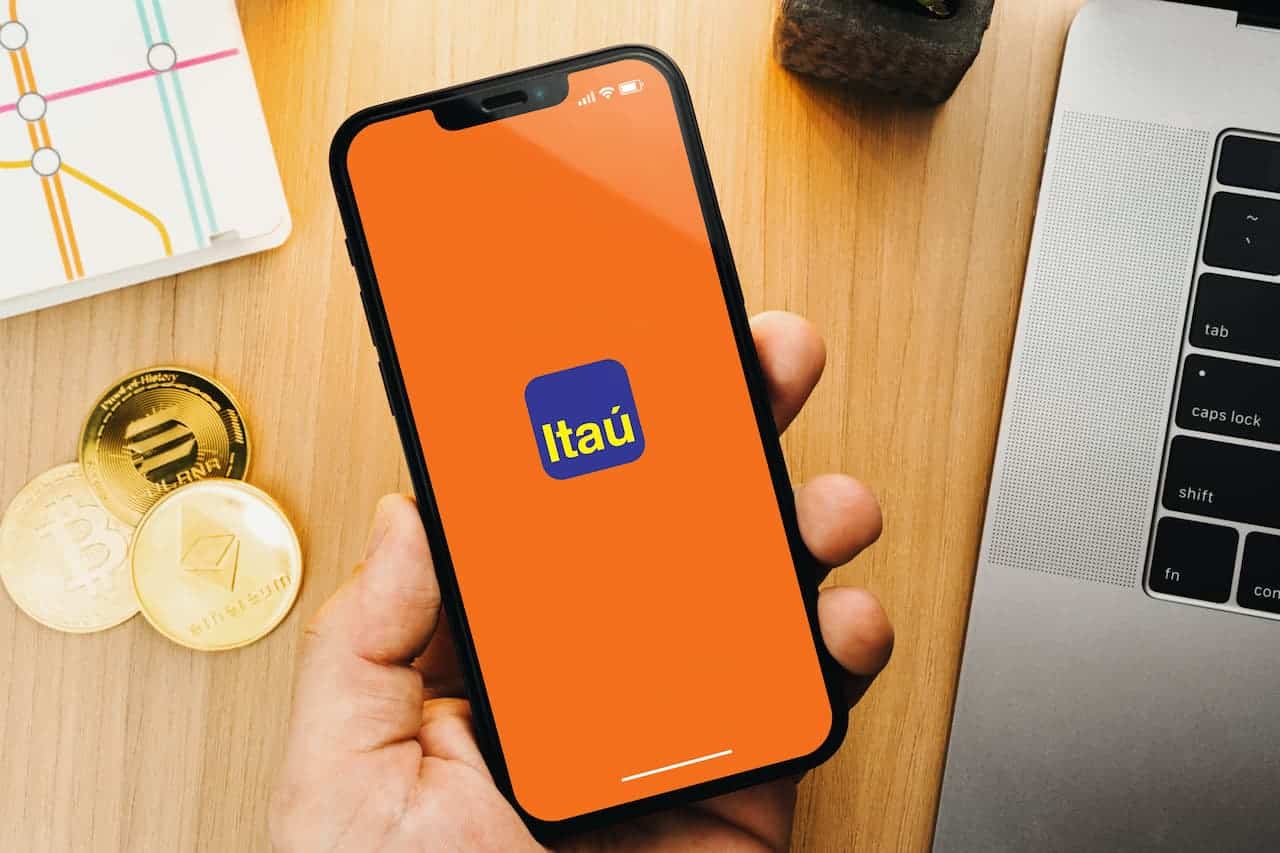 Mão segurando celular com logo do Itaú. Criptomoedas em bancada