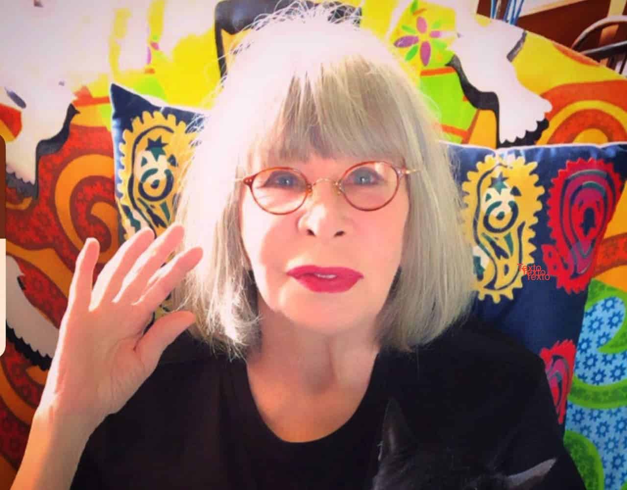 A cantora Rita Lee de cabelos brancos e óculos, usando uma camiseta preta e acenando para a câmera