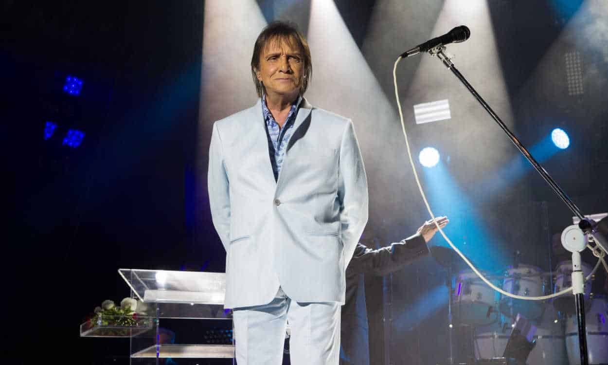 O cantor Roberto Carlos usando um terno branco durante show, ao lado de um microfone pedestal.